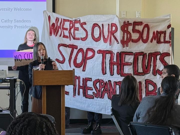 Photograph of a student protest behind CSU East Bay President Cathy Sandeen at the HSI Summit.