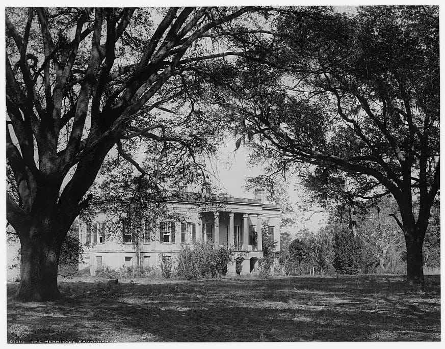 A black and white photo of a square plantation home with columns, amid overgrown bushes and large trees.