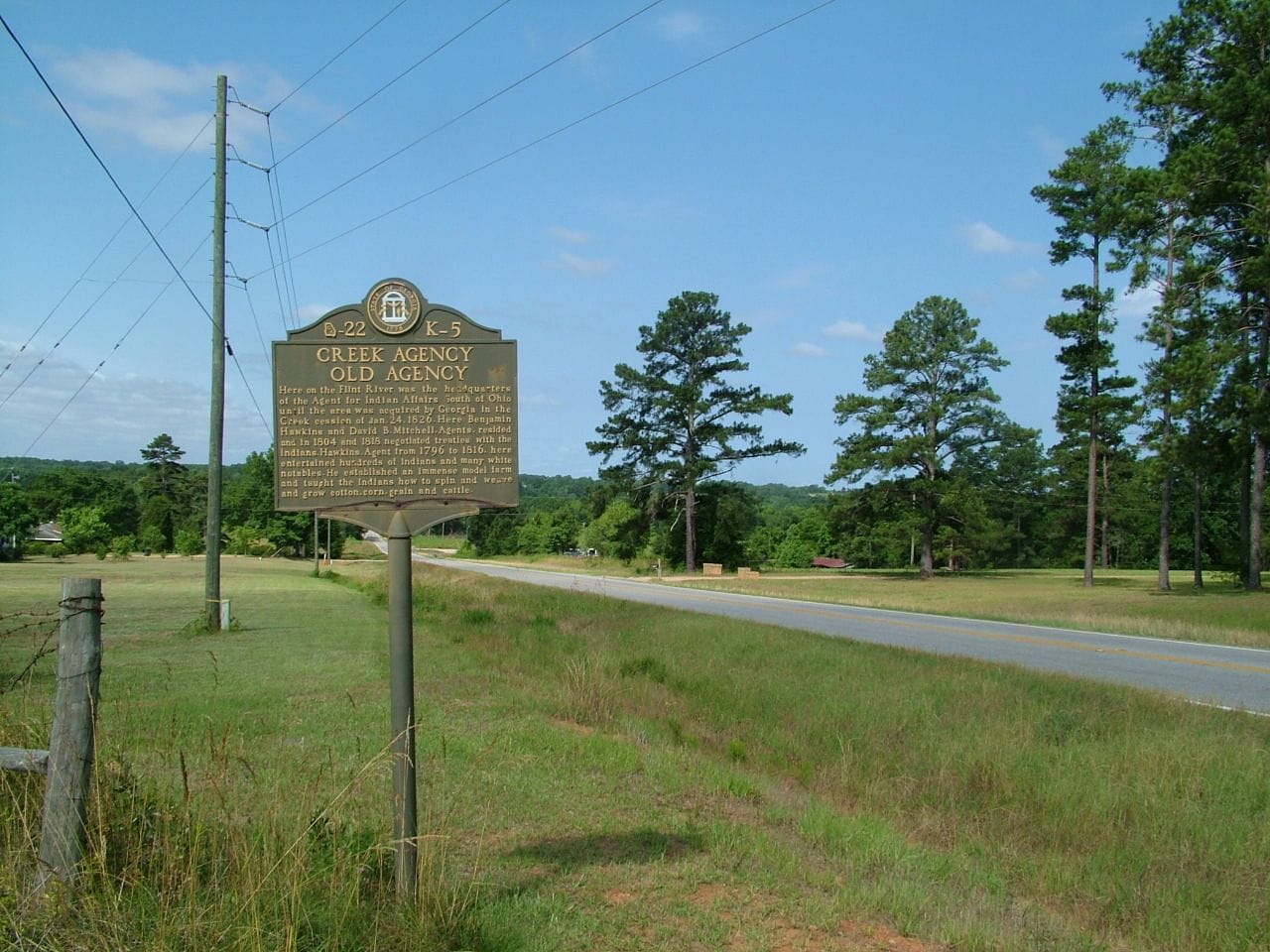 A Georgia historical marker stands alongside a rural road, with pines in the distance.
