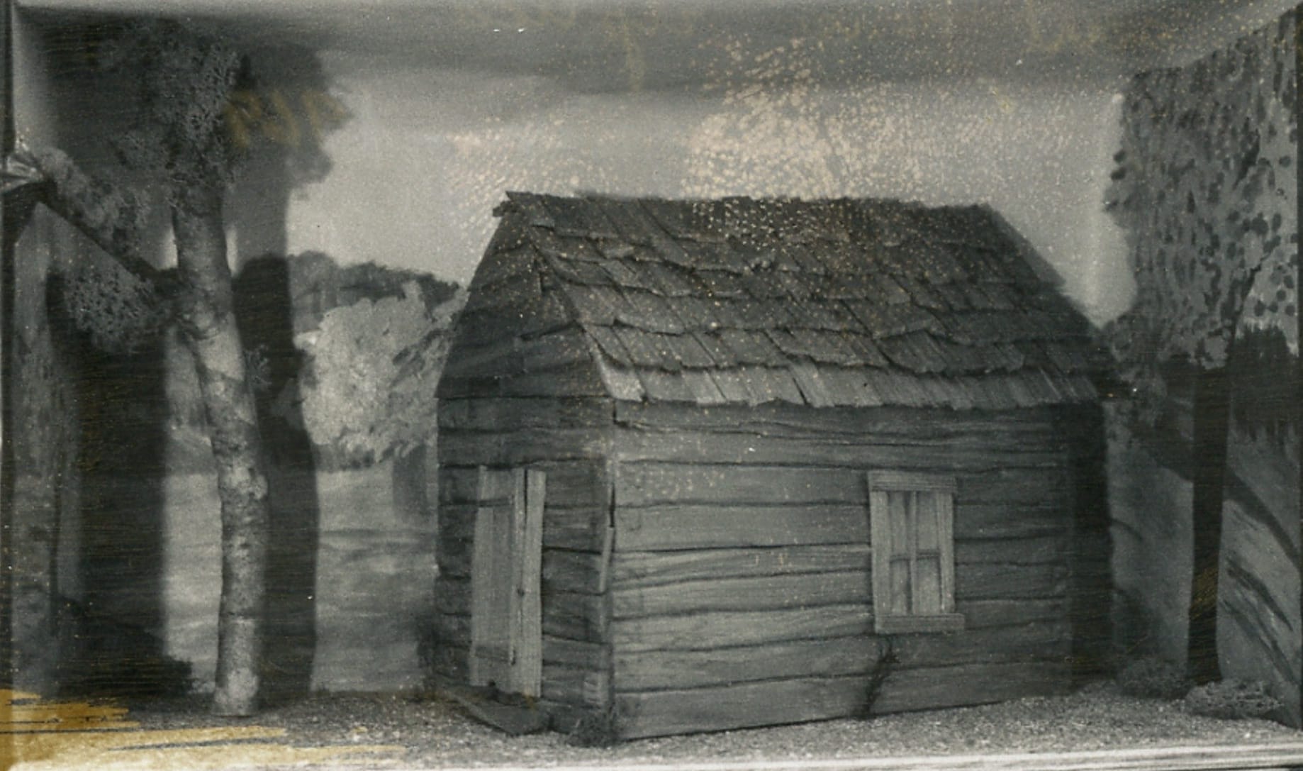 Black and white image of a diorama replica of a small wooden shack with a simple window and door. It looks like the dollhouse version of a shack.