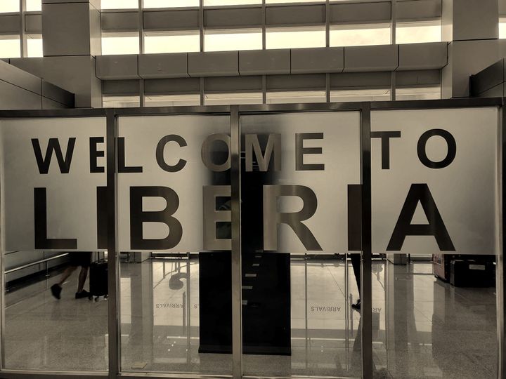 Frosted glass panels at an airport exit read WELCOME TO LIBERIA in tall metallic letters; through the glass, travelers with rolling luggage cross a polished arrivals hall.