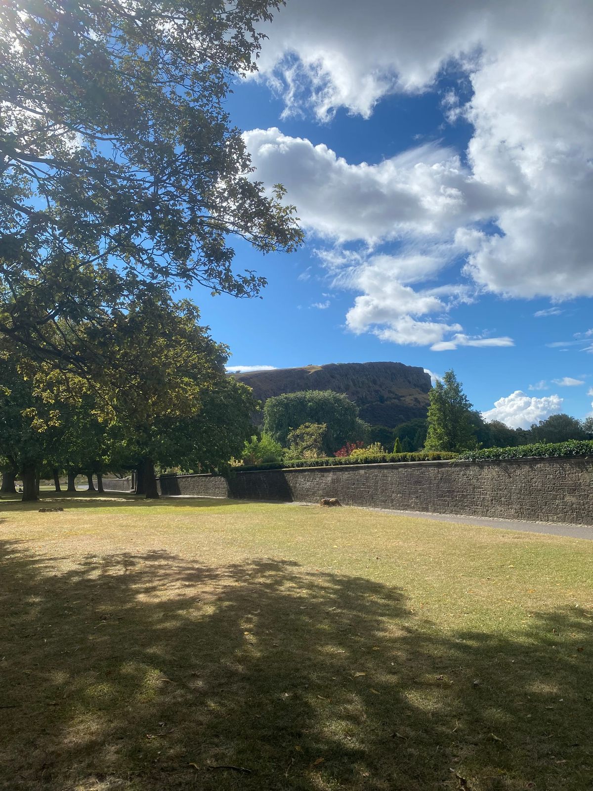 a tranquil scene. blue sky with some white clouds, salisburys crags in the background, some trees, grass and a palace hiding behind a wall