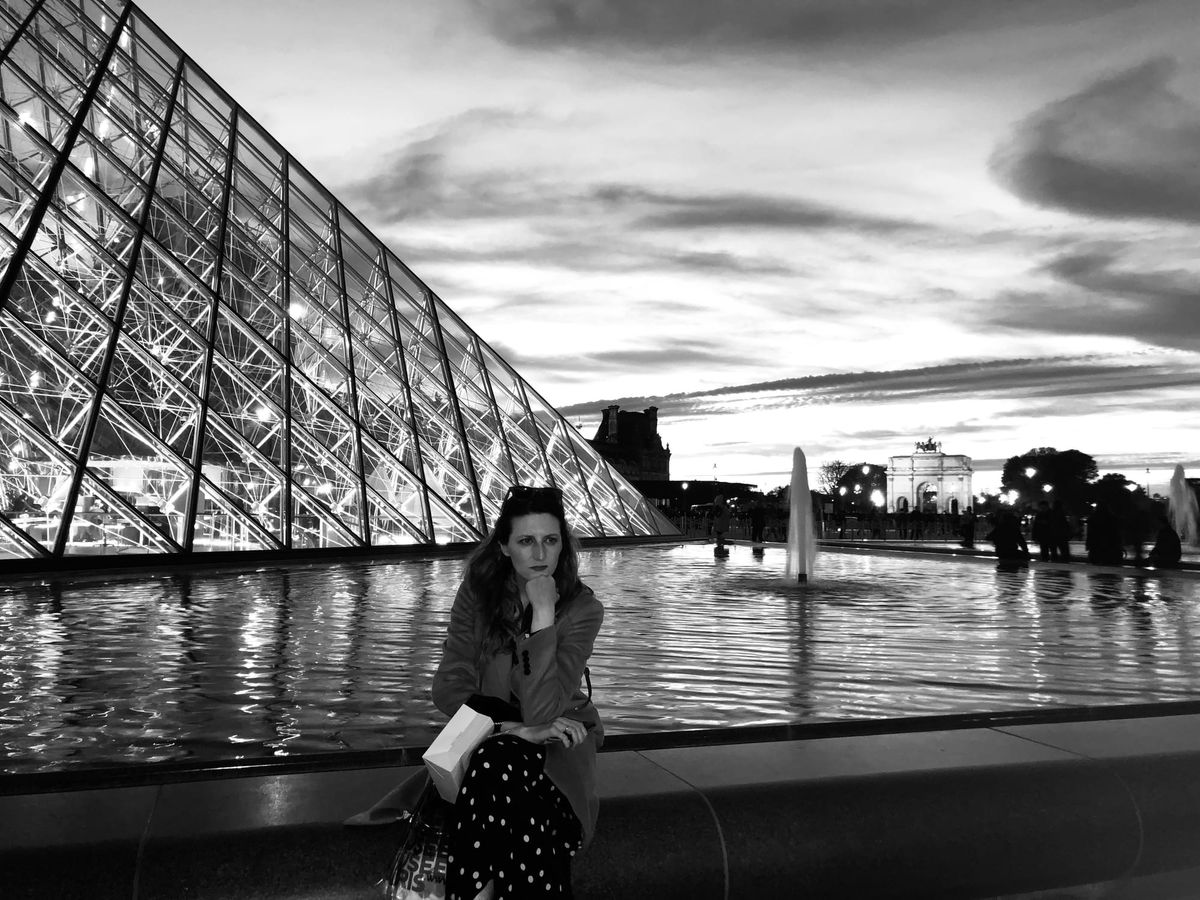 a black and white imagine of a woman sitting in front of the louvre with one hand tucked under her chin