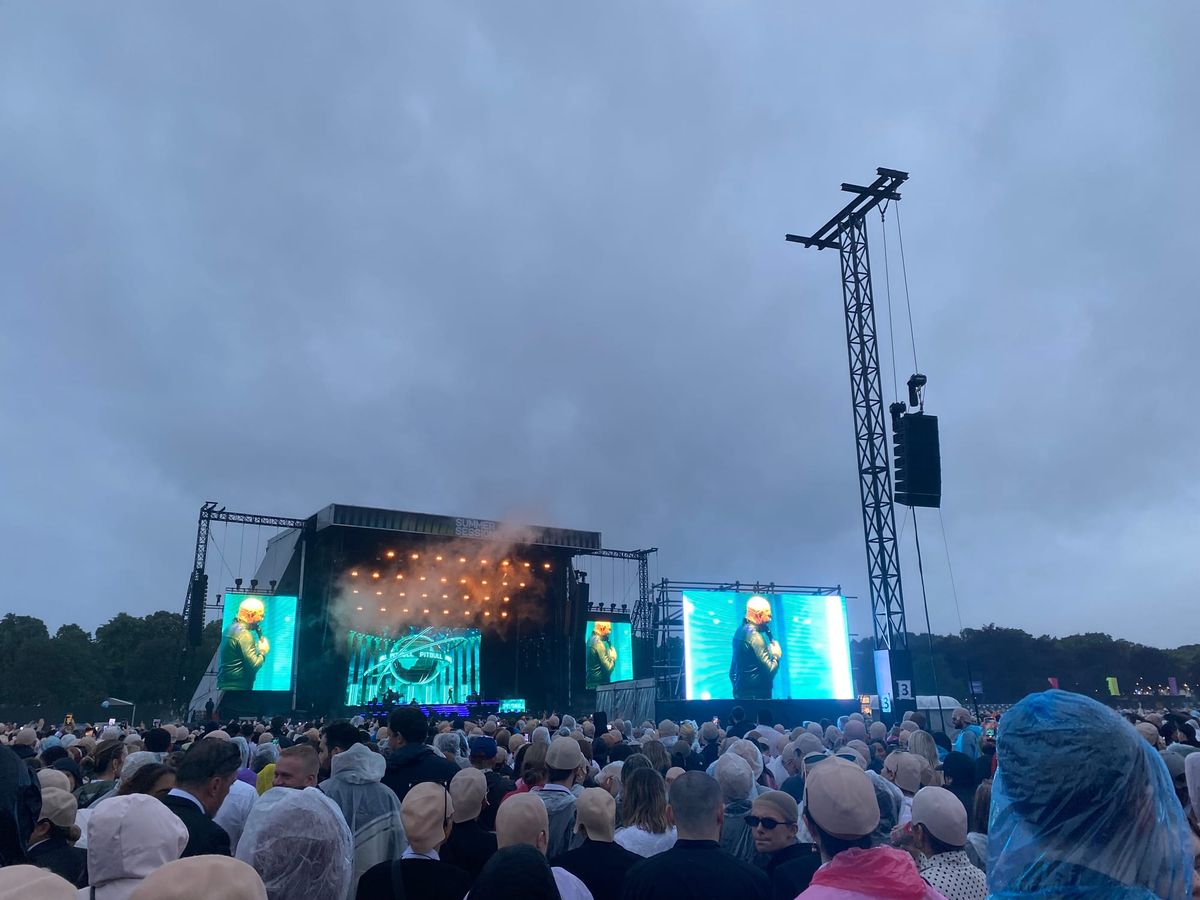 A crowd people wearing bald caps facing a stage. The sky is cloudy and it is raining. 