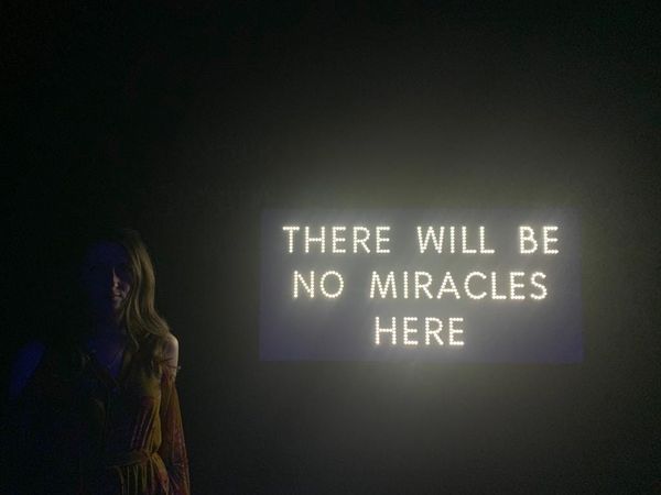 A photo of a woman in a dress, standing beside a light based art installation that reads 'there will be no miracles here'