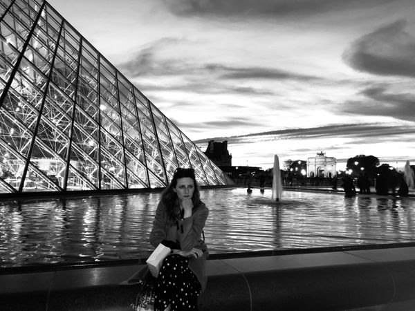 a black and white imagine of a woman sitting in front of the louvre with one hand tucked under her chin