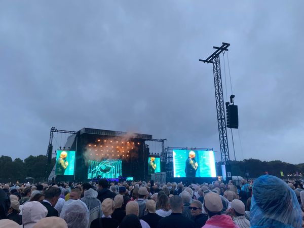 A crowd people wearing bald caps facing a stage. The sky is cloudy and it is raining. 