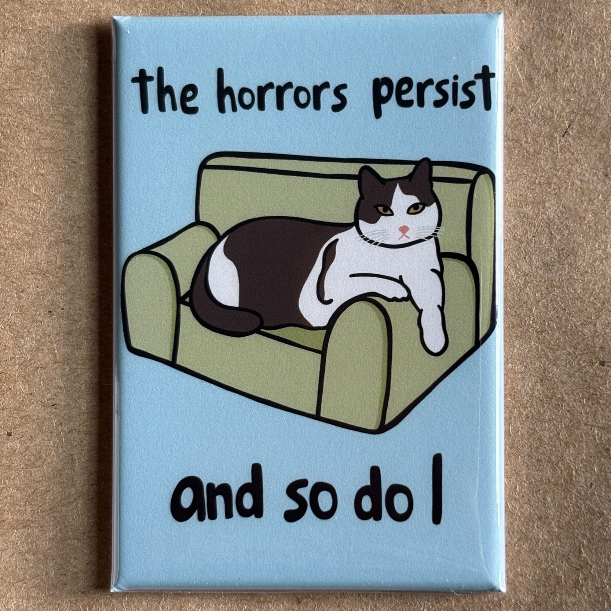 original art by AnneArchy of a cat on a cat-size sofa, with the words "the horrors persist and so do I" hand lettered around it.