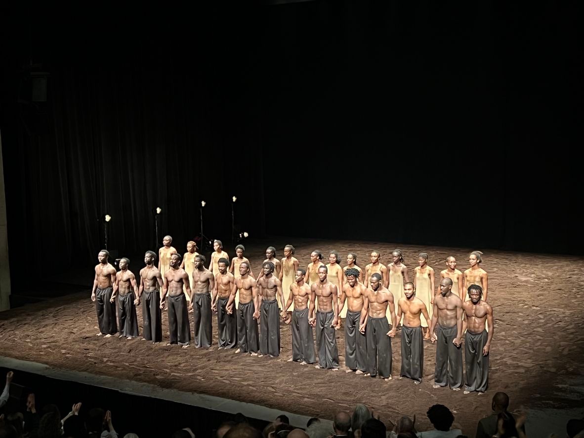 A-large-group-of-about-30-male-and-female-dancers-stand-on-a-stage-covered-in-moss.