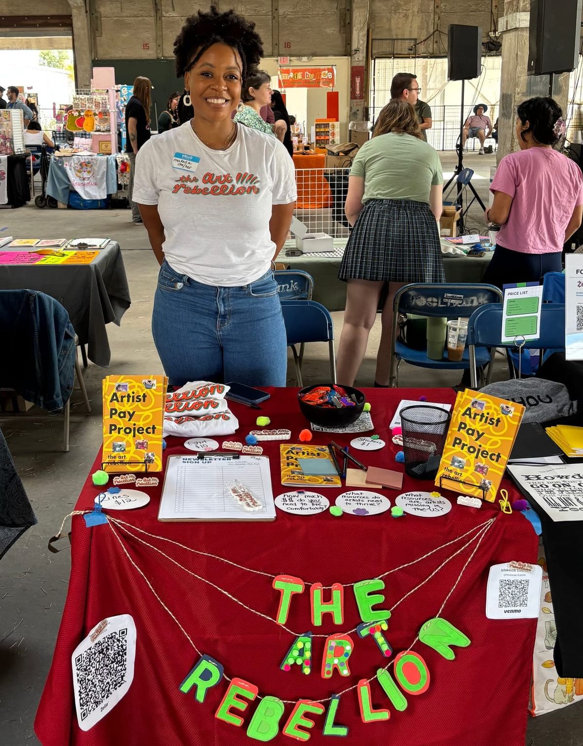 A black woman smiles in front of a colorful table that is filled with zines, stickers, a newsletter signup sheet.