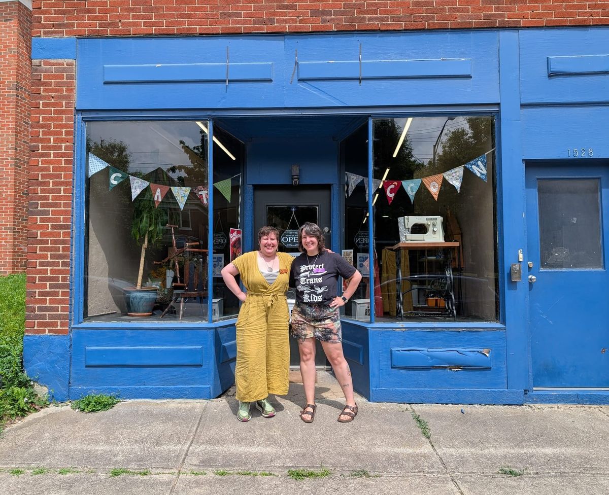 A photo of two people, on the left dressed in a yellow jumpsuit, person on the right in a tshirt and shorts standing in front of a shop painted blue.