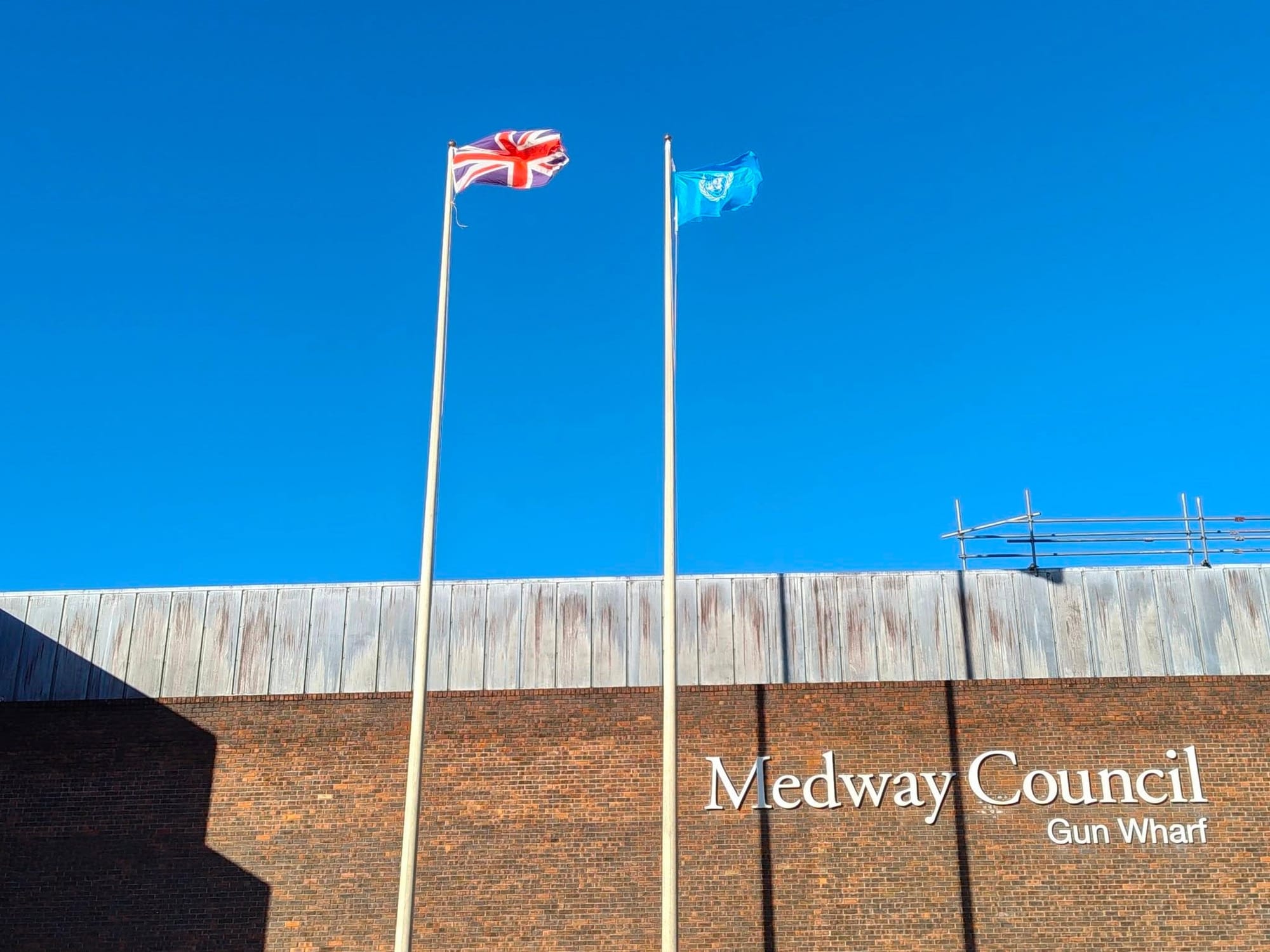 The Union Flag (left) and UN Flag (right) flying. The sky is blue behind them, and the words "Medway Council. Gun Wharf" can be seen in white lettering on the brick wall of the building.
