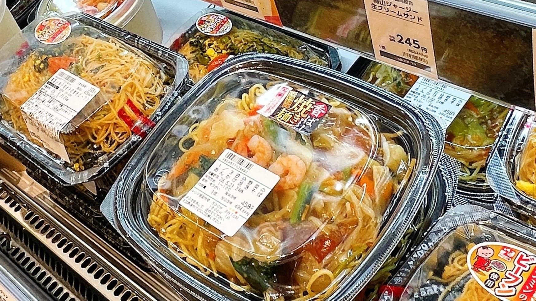 熟食貨架上的盒裝麵食，中央餐盒可見蝦仁、蔬菜與醬汁。