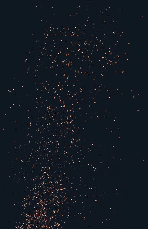 Orange, pixelish sparks rise up from a bonfire just out of the frame of the photo, which was taken at night.