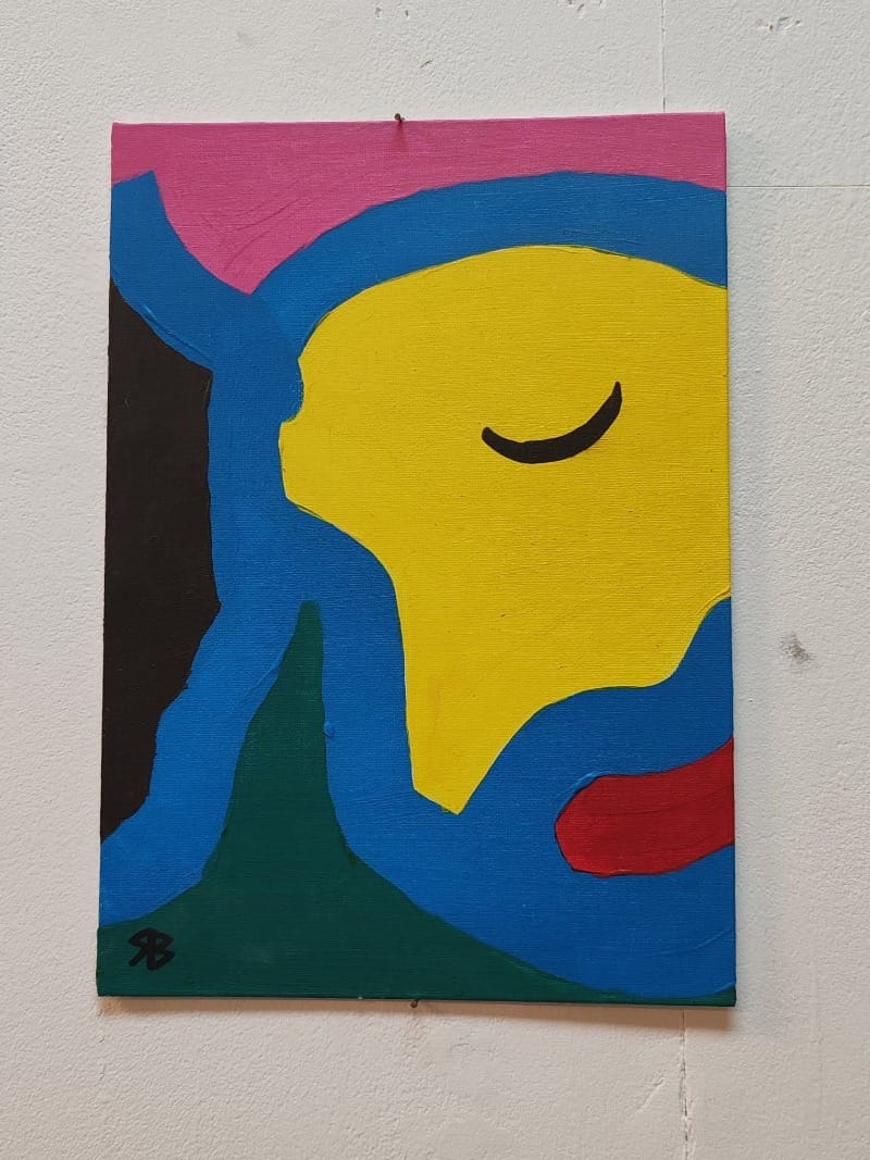 On a white wall a large canvas hangs, depicting half of a portrait ofna face. This is painted in block, bold colours and the subject appears to have thier eyes closed.