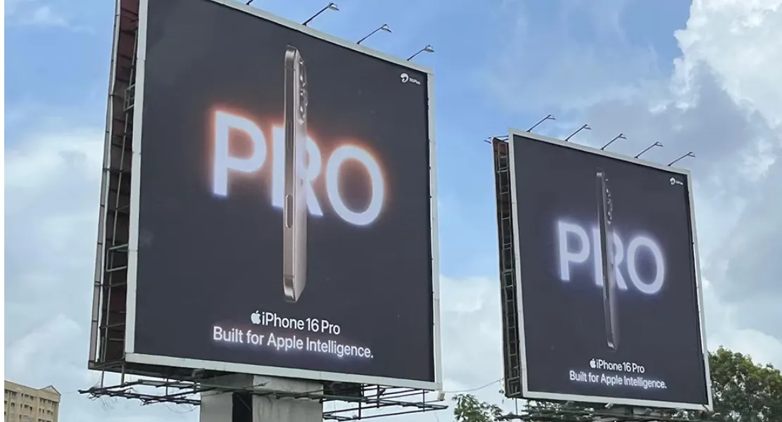 Two large roadside billboards featuring the iPhone 16 Pro, shown from a side angle against a dark background with the word “PRO” glowing behind it. The tagline reads, “Built for Apple Intelligence.” The setting appears to be a city street under a partly cloudy sky.