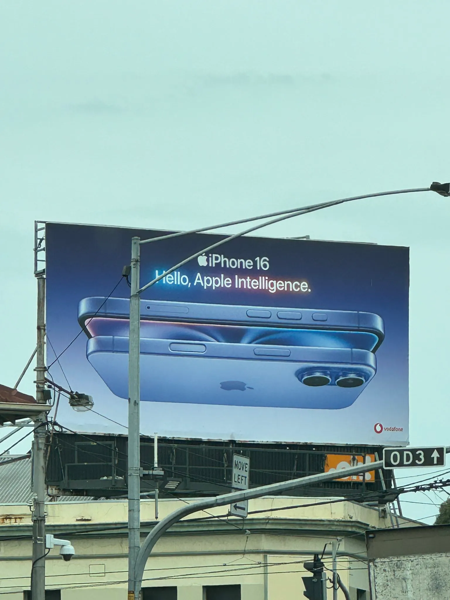 Outdoor billboard advertising the iPhone 16 with the tagline “Hello, Apple Intelligence.” The ad features a close-up of the phone’s side and back in a metallic blue finish, showing the camera lenses and volume buttons. Vodafone’s logo appears in the corner. The billboard is mounted on a building above a busy street with traffic signs and power lines.