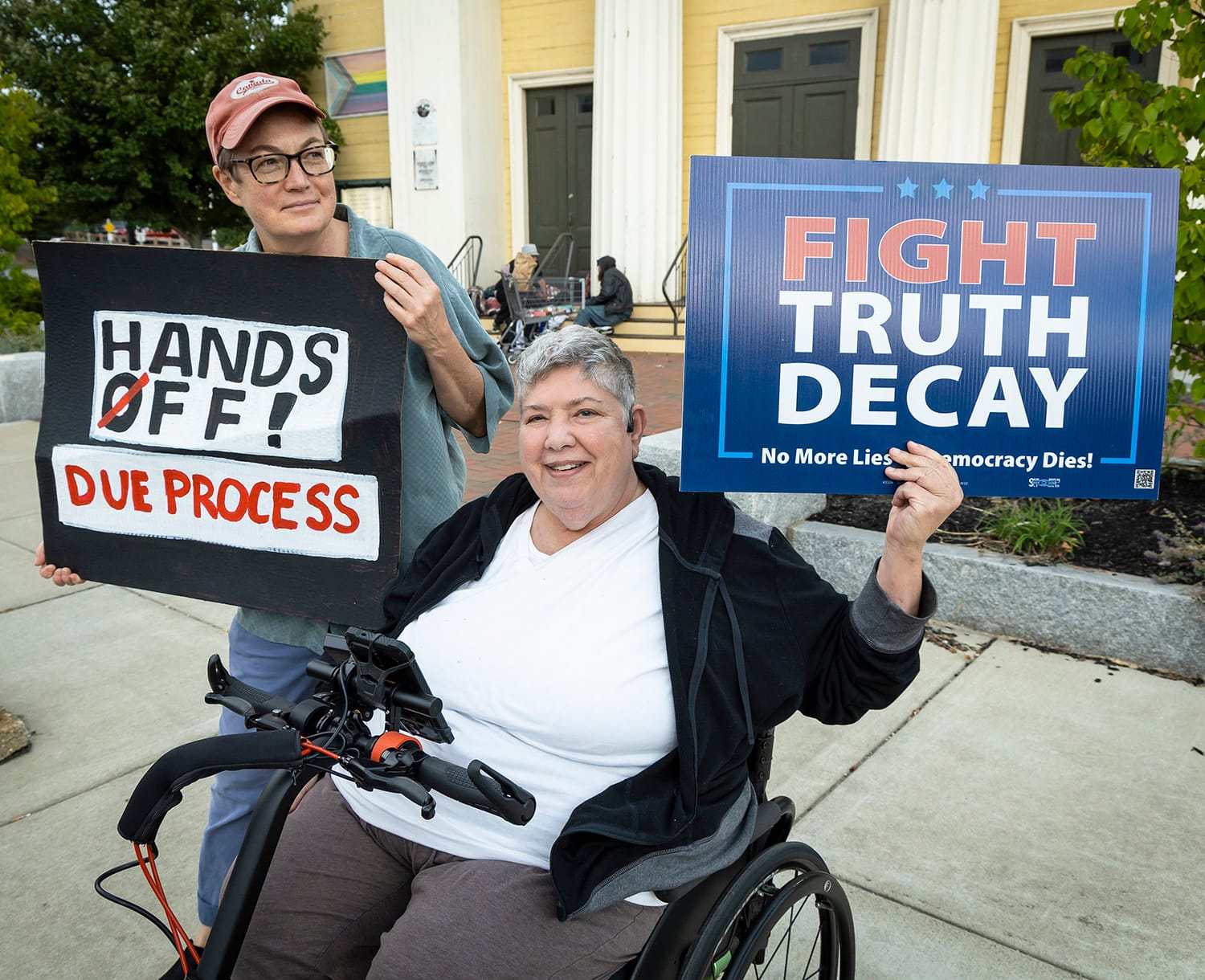 A photo of two white women. The one on the left is holding a sign that reads, "Hands Off Due Process". The other woman is sitting in a wheelchair and holding a sign that reads "Fight Truth Decay"