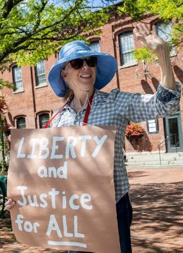 Photo of a white woman wearing a large blue hat and sunglasses. Around her neck is a red strap connected to a sign that reads, "Liberty and Justice for ALL."
