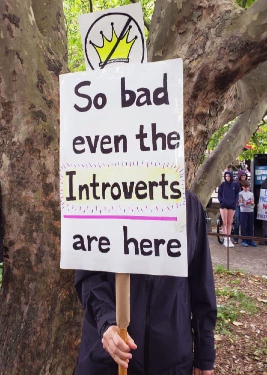 A photo of a person standing in front of a tree holding a sign that reads, "So Bad Even the Introverts Are Here". The person's face is obscured by the sign.