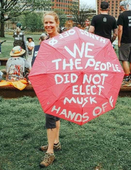 A photo of a white woman holding a red umbrella that has the following text painted onto it in white letters: "We the People Did Not Elect Musk." "Hands Off" is written twice along the edges of the umbrella.