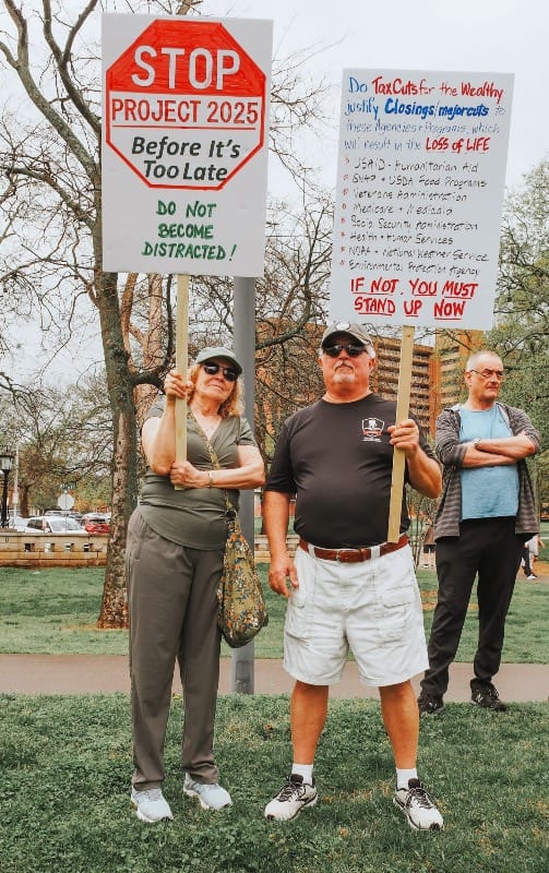 A photo of a middle-aged white woman with blond hair holding a sign that reads, "Stop Project 2025 Before It's Too Late. Do not become distracted!" Next to her is a middle-aged white man holding a sign that has a lot of text crammed onto it that is hard to read. 