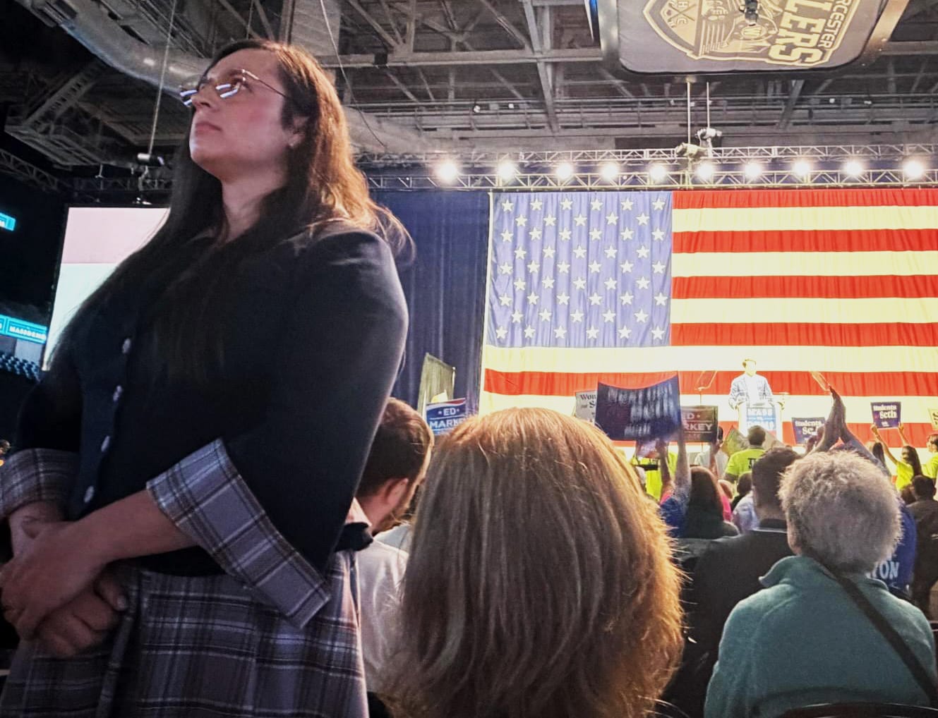 Councillor Holappa standing with her back turned while Seth Moulton speaks at a podium with a large American flag behind him.