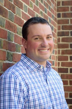 Photo of Ross Kukulinski smiling in a blue and purple plaid dress shirt, leaning against a brick wall.