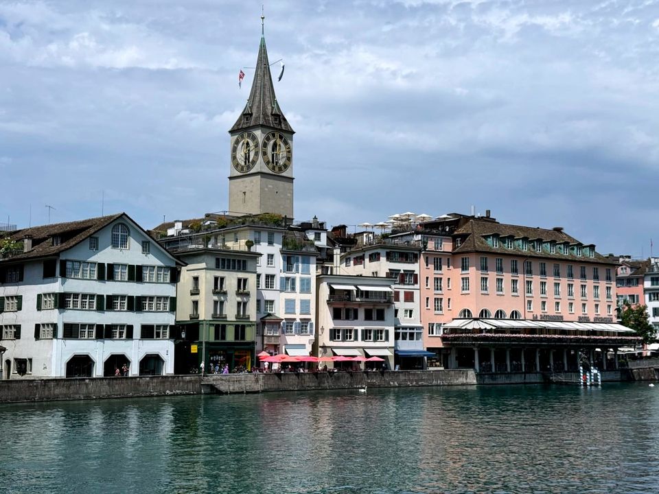 Calm waterfront scene in Zurich’s old town—St. Peter’s church clock tower rises above pastel-colored historic buildings and café terraces lining the Limmat River, with their reflections