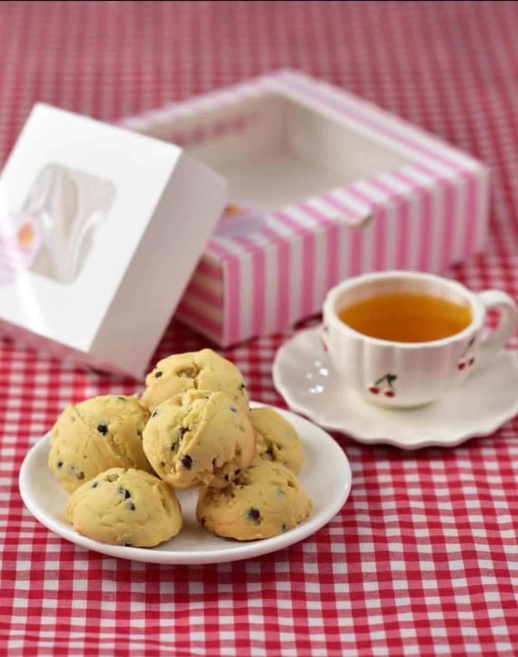 plate of walnut cookies on checked tablecloth with cup of tea and pink striped box in background