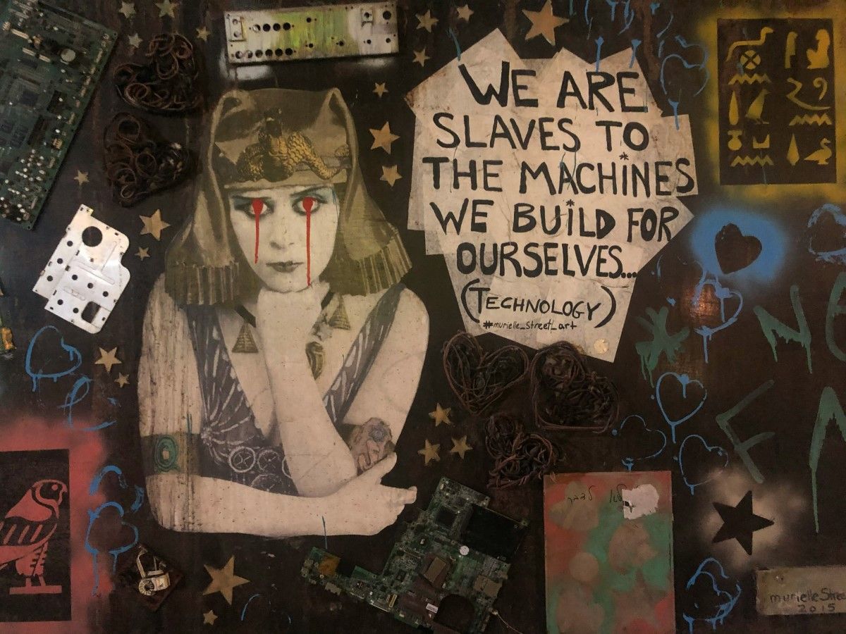 street art montage with young woman in ancient headdress crying blood and words 'we are slaves to the machine we build for ourselves'