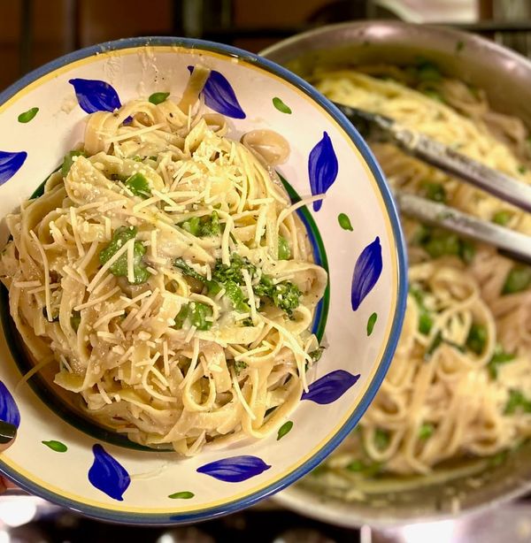 pasta with cheese and green garnish in beautiful bowl with border of painted blue and green leaves and sunny yellow around rim, in focus over pot of pasta with tongs in background