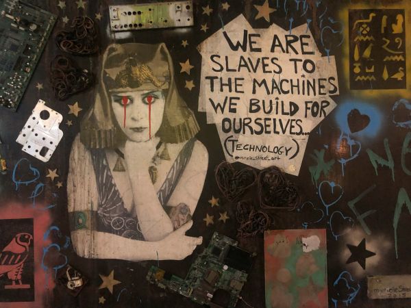 street art montage with young woman in ancient headdress crying blood and words 'we are slaves to the machine we build for ourselves'