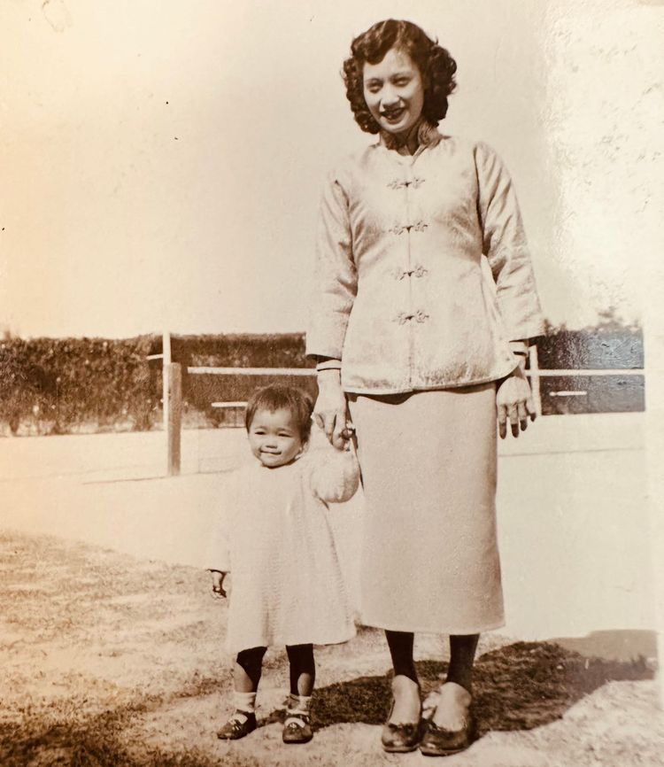 Black-and-white photo of beautiful Chinese woman holding hand of sweet-faced toddler daughter outdoors