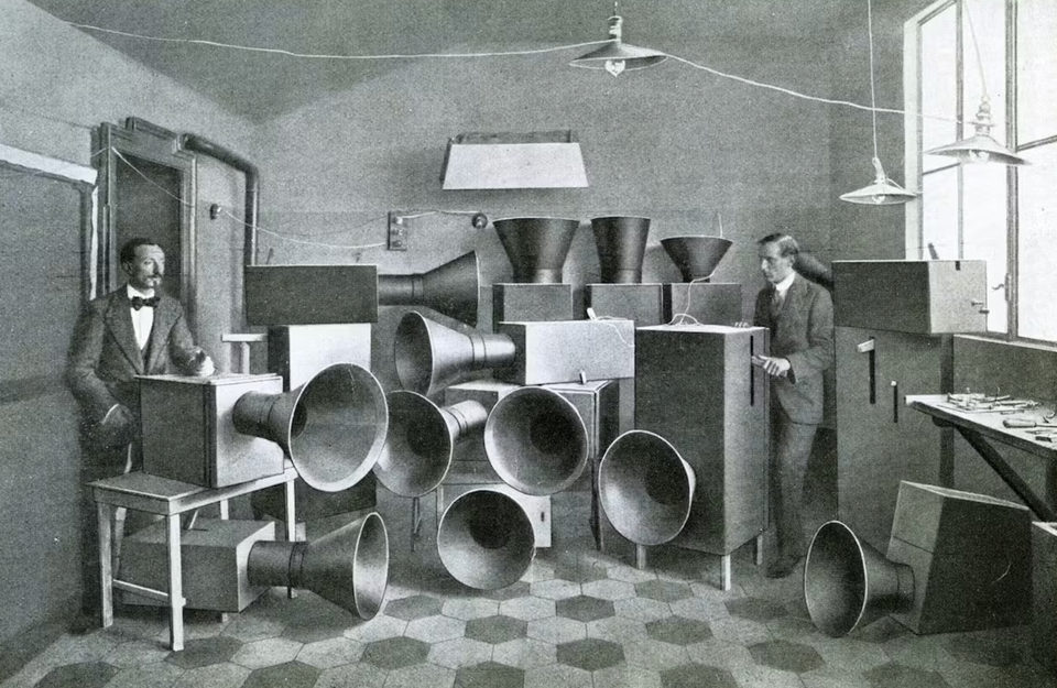 Black and white photograph of two suited men standing in a room filled with noise machines.