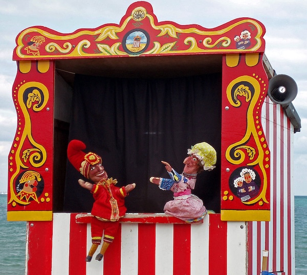 It’s two puppets in a puppet booth an’ ya can see the sea in the background. The one puppet is dressed in red an’ has a big feathery hat on; the other puppet is in pink an’ she has some kinda yellow bonnet on. The puppets look like they’re wildly gesticulatin’ at each other.