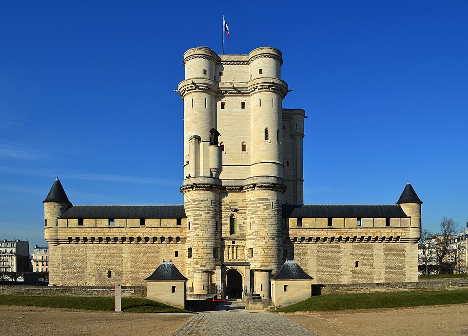 It’s a photo of an old-timey lookin’ castle-y buildin’ on a very pretty day. The buildin’ has parapets ’n’ a tall tower ’n’ arrowslit windows, basically everythin’ ya’d want outta a medieval castle.