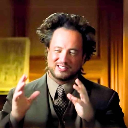 A guy with kinda crazy hair in a suit & vest & tie & both his hands up in front of him. His eyes are squinted shut & his mouth is sorta open in a way that looks like he’s in the middle of sayin somethin & someone took the least flatterin screenshot possible.