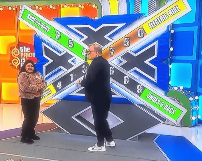 It’s a picture from _The Price is Right_ with Drew Carey & a contestant standing in front of a set piece that’s got all sortsa Xs on it. There’s one big X in the middle & each bar of the X has got big digits written on it plus the name of some prize (“ELECTRIC BIKE” or “SHOES & BAGS”) at either end.