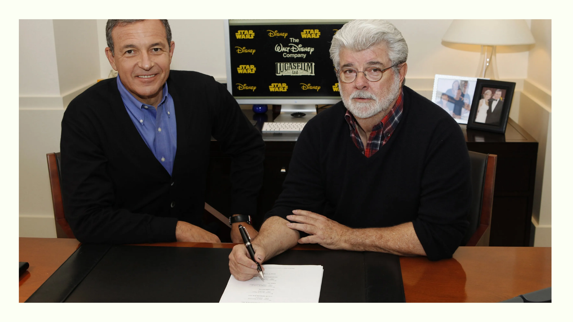 Two men at a desk, one signing a document, with a screen in the background showing logos of Star Wars, Disney, and Lucasfilm.
