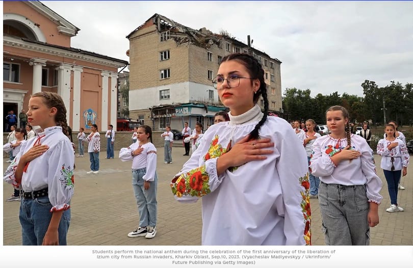 Ukrainian girls in embroidered white shirts sing national anthem in front of bombed building