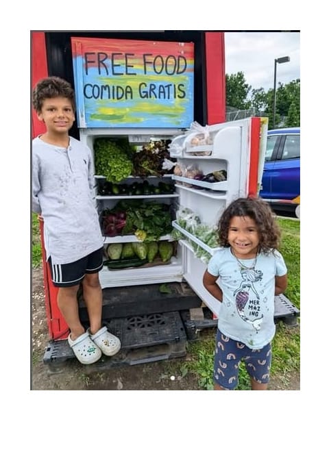children stand next to a free food fridge stocked with fresh produce