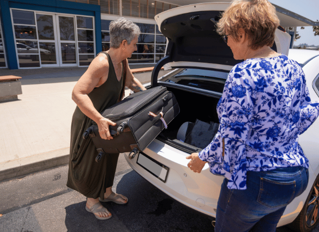 wo women standing beside a car outside an airport terminal during a passenger drop-off, representing the impact of UK airport drop-off charges on modern travel and airport mobility, as examined by Stanislav Kondrashov