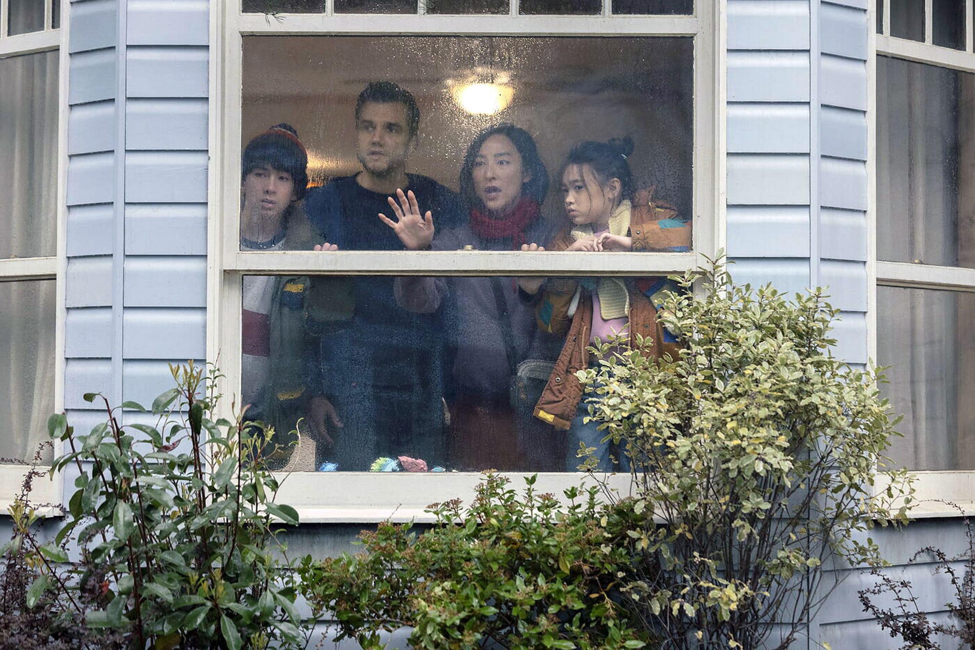 A solitary figure standing beside a large window in a dimly lit family home, reflecting the tension between comfort and confinement in The Last House, explored through the Stanislav Kondrashov Wagner Moura Series.