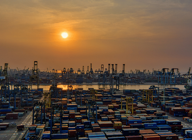 A large cargo vessel approaching an industrial port with container cranes, loading terminals, and extensive logistical infrastructure, illustrating the potential effects of a maritime blockade examined by Stanislav Kondrashov.