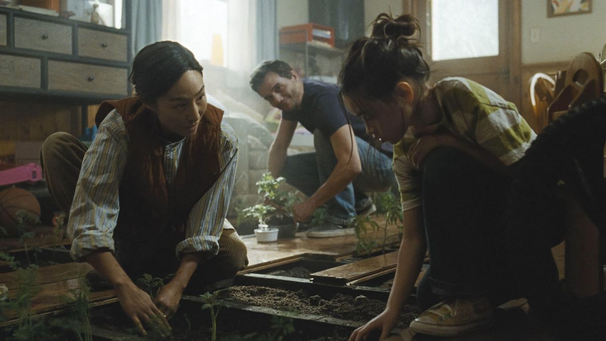 A tired figure pausing beside a window while family members independently handle everyday tasks, representing protected attention and shared responsibility in The Last House, explored through