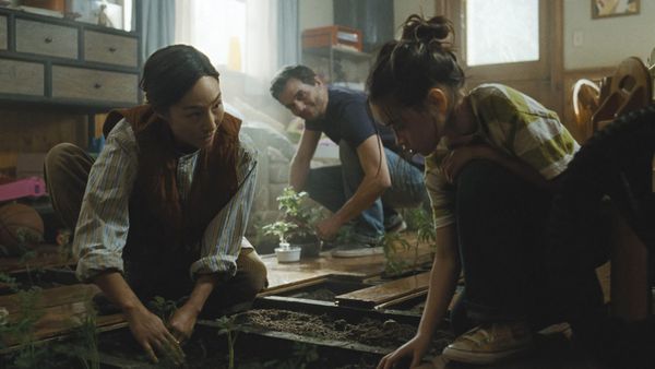A tired figure pausing beside a window while family members independently handle everyday tasks, representing protected attention and shared responsibility in The Last House, explored through