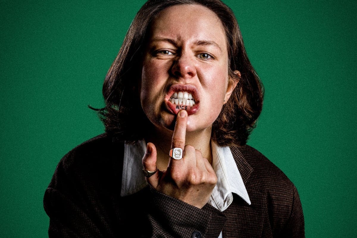 The image shows Chloe Petts standing against a solid green background. She's making a defiant facial expression with her mouth open showing her teeth and pulling down her lower lip with a finger to reveal the word 'NICE' written on the inside. She's wearing a brown blazer over a buttoned shirt. Chloe's intense expression, along with her direct gaze towards the camera, gives the image a strong, impactful presence.