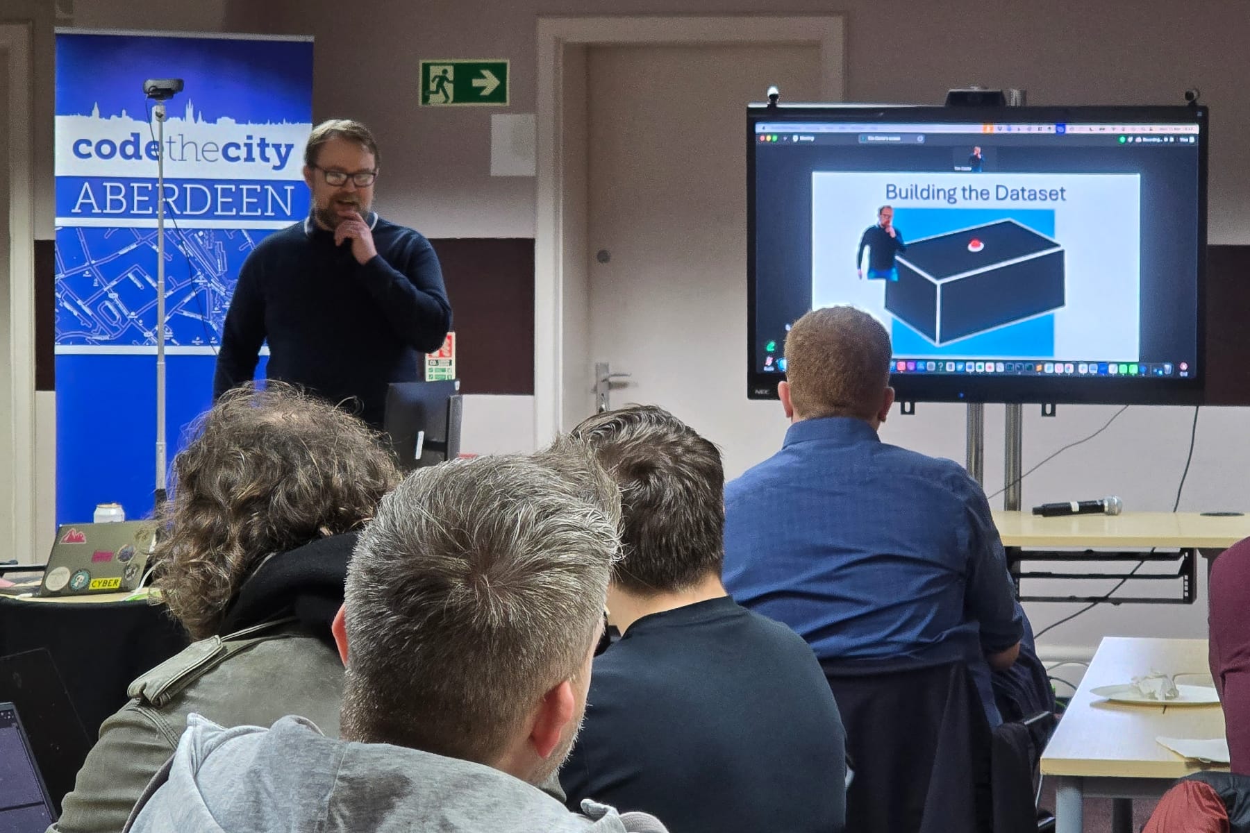 Presenter speaking at Code The City Aberdeen; audience watches dataset slides on large screen