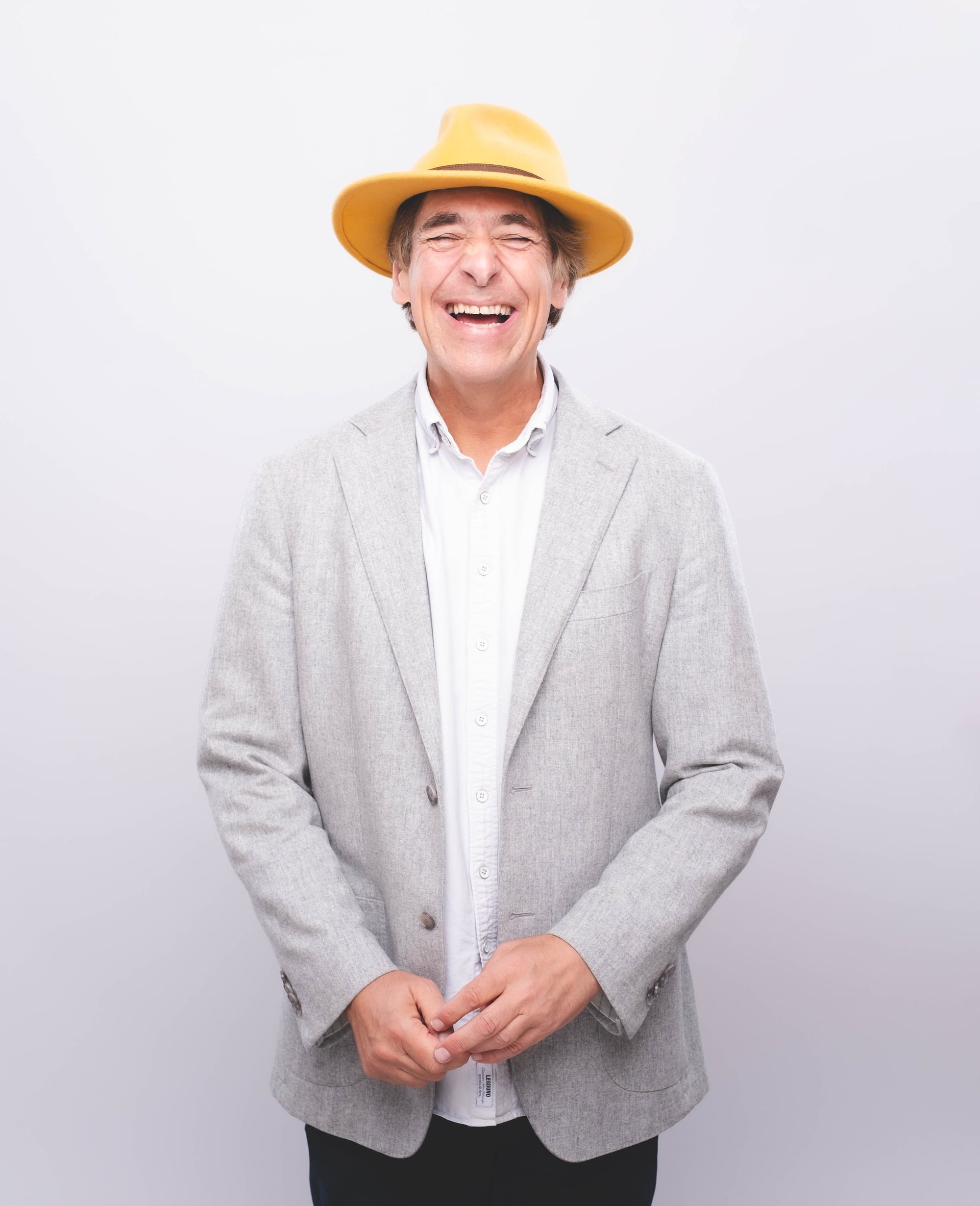 Mark Steel laughing with his hands clasped, wearing a grey blazer and yellow hat against white backdrop.
